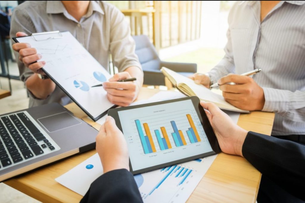 Three professionals in a modern office collaborating over data. One holds a tablet with a bar chart, another points to a clipboard with line graphs, and a third takes notes in a journal.